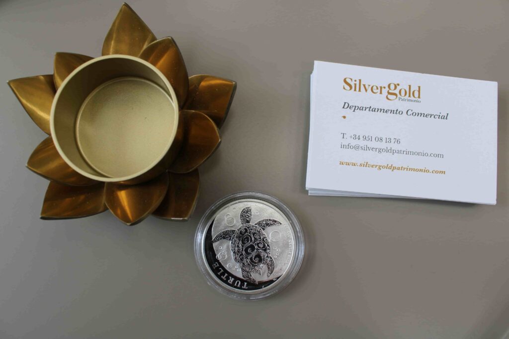 Una moneda de plata de la Tortuga reposa en una mesa junto a una tarjeta de SilverGold Patrimonio